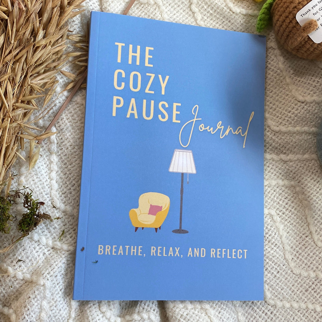 A blue hardcover journal titled 'The Cozy Pause Journal' with gold lettering, styled on a white textured knit blanket with dried wheat, a crocheted sunflower, a small plush character, and a blue mug.