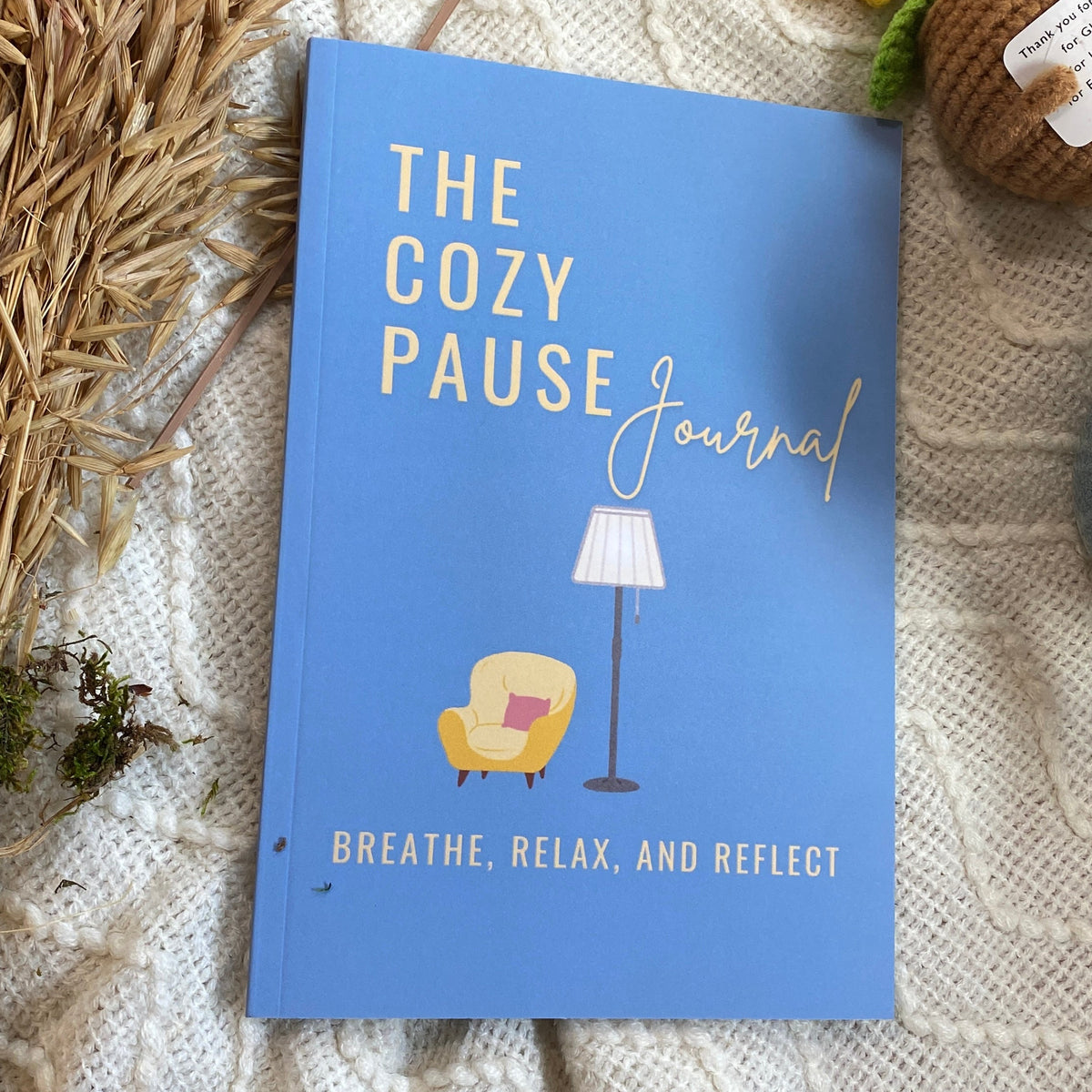 A blue hardcover journal titled 'The Cozy Pause Journal' with gold lettering, styled on a white textured knit blanket with dried wheat, a crocheted sunflower, a small plush character, and a blue mug.